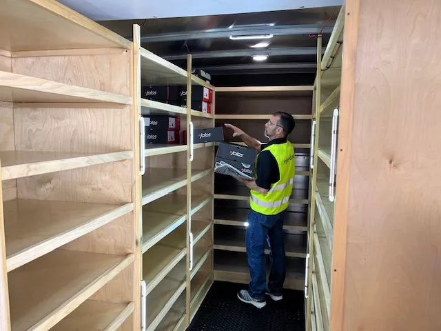 Man wearing a high-visibility vest stocking Jalas shoe boxes on shelves inside a mobile shoe truck.