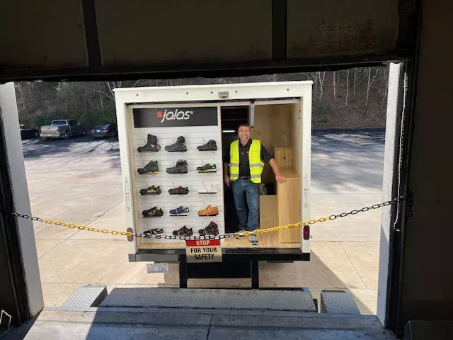 Man wearing a high-visibility vest standing in the back of a Jalas safety shoe display truck with shelves of work shoes.